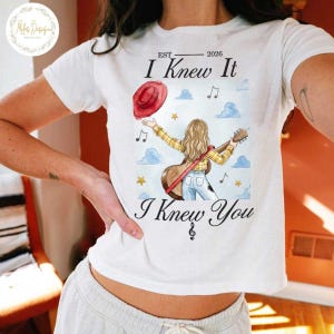 Op de afbeelding: Een wit t-shirt met een afbeelding van een persoon met lang blond haar in een geel geruit overhemd en een blauwe spijkerbroek, die een gitaar vasthoudt. Boven en onder de illustratie staat de tekst "EST 2026 I Knew It I Knew You" met muzieknoten.
