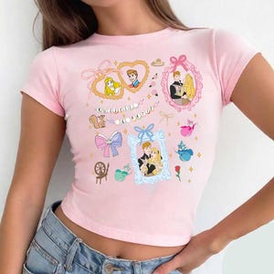 Op de afbeelding: Een roze crop top met korte mouwen met een grafische print van Doornroosje en Prins Philip in verschillende frames en illustraties. De tekst "KUTRELL & KATIE" is op het shirt gedrukt.