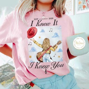 Op de afbeelding: Een lichtroze t-shirt met een afbeelding van een persoon met lang blond haar in een geel geruit overhemd en blauwe spijkerbroek, met een gitaar. De tekst luidt EST 2026 I Knew It I Knew You, omringd door rode hoeden, sterren en muzieknoten.