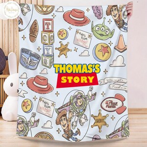 May include: A light blue blanket featuring cartoon characters and elements from the movie "Toy Story." The design includes Woody, Buzz Lightyear, and other iconic imagery, with the text "THOMAS'S STORY" in bold letters. The blanket has a playful and colorful design.