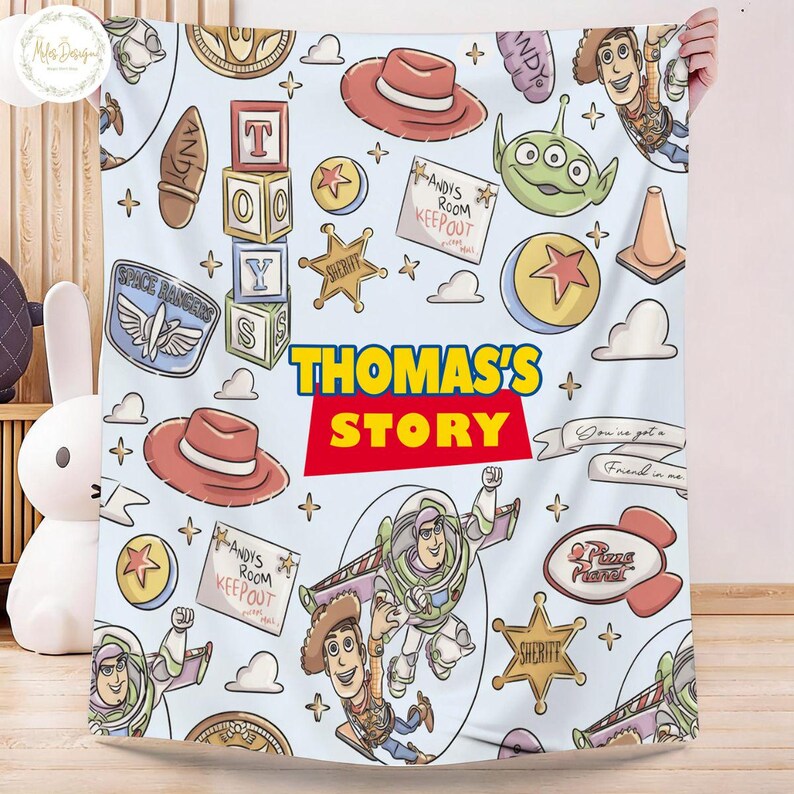 May include: A light blue blanket featuring cartoon characters and elements from the movie "Toy Story." The design includes Woody, Buzz Lightyear, and other iconic imagery, with the text "THOMAS'S STORY" in bold letters. The blanket has a playful and colorful design.