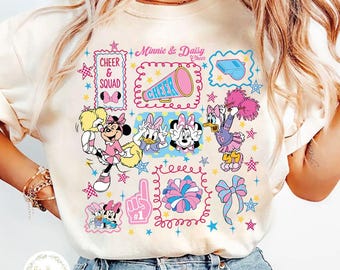 May include: Cream-colored t-shirt featuring Minnie and Daisy Duck cheerleading graphics. The design includes the text "Minnie & Daisy Cheer" and "Cheer & Squad", along with cheerleading elements in pink, blue, and yellow.