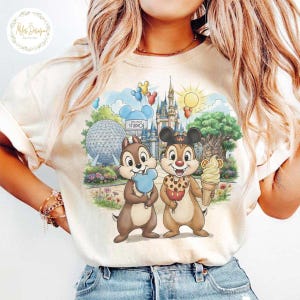 May include: Cream-colored graphic tee featuring Chip and Dale holding ice cream cones in front of a Disney park scene with a castle, Spaceship Earth, and the Hollywood Studio sign. The shirt has a round neckline and short sleeves.