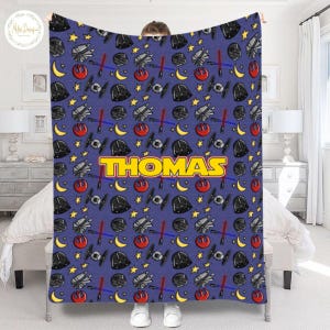 May include: A blue blanket featuring a repeating pattern of stars, moons, spaceships, and Darth Vader helmets. The name "THOMAS" is displayed in large yellow letters. The blanket is held up by a person.