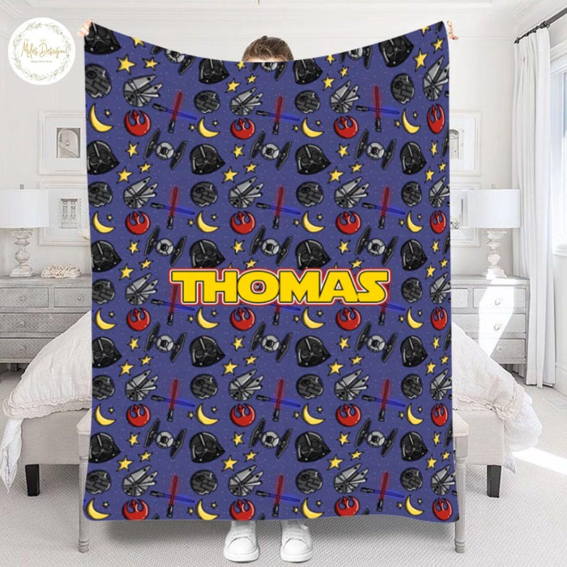 May include: A blue blanket featuring a repeating pattern of stars, moons, spaceships, and Darth Vader helmets. The name "THOMAS" is displayed in large yellow letters. The blanket is held up by a person.