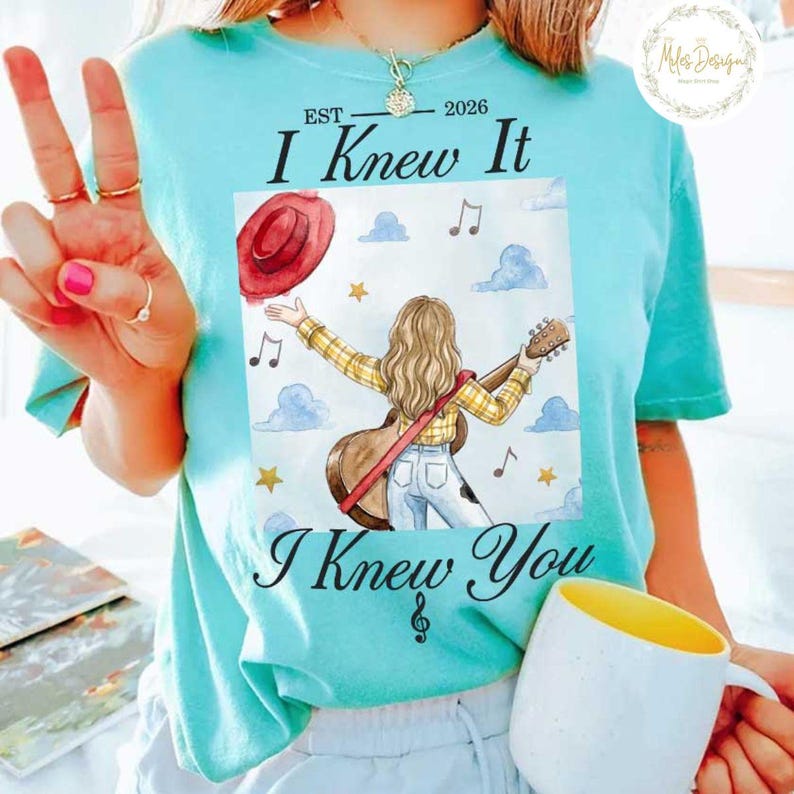 Op de afbeelding: Een licht turquoise t-shirt met een afbeelding van een persoon met lang haar die gitaar speelt, gekleed in een geel geruit overhemd en een blauwe spijkerbroek. De tekst luidt EST 2026 I Knew It I Knew You, omringd door wolken, sterren en muzieknoten.