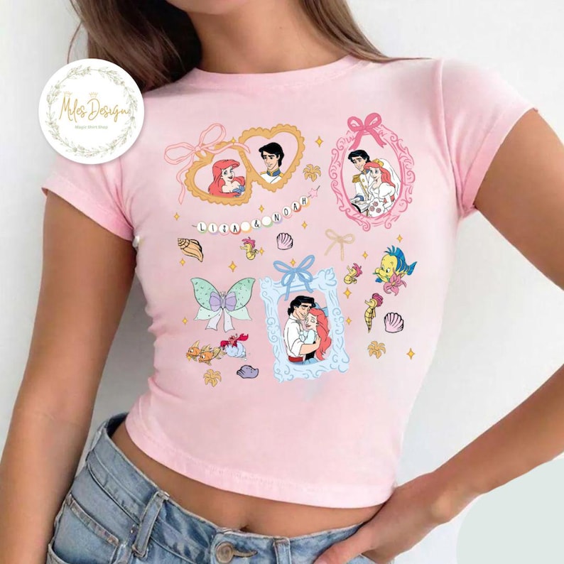 Puede incluir: Camiseta rosa de manga corta y corte crop con un estampado gr&aacute;fico de Ariel y el Pr&iacute;ncipe Eric de La Sirenita. El estampado incluye varias im&aacute;genes de la pareja en diferentes poses, junto con conchas marinas, estrellas de mar y una mariposa.