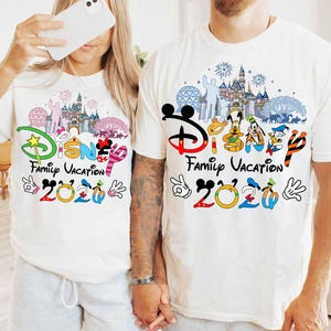 May include: Two white t-shirts with a colorful Disney Family Vacation 2020 design. The design includes Disney characters, a castle, fireworks, and the Disney logo. The shirts are short-sleeved.