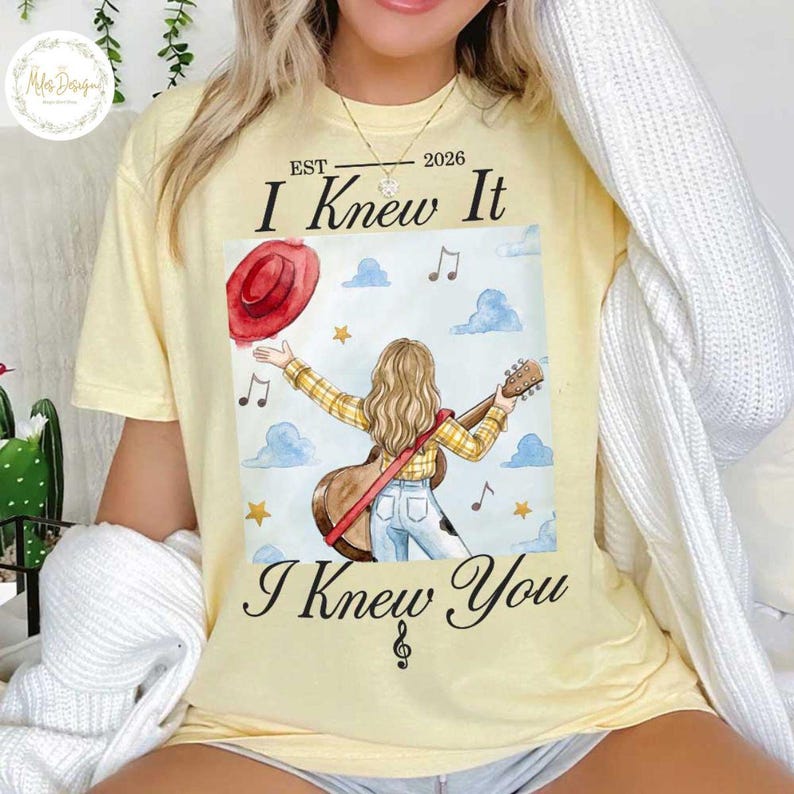Op de afbeelding: Een lichtgeel t-shirt met een afbeelding van een persoon met lang haar in een geel geruit overhemd en een blauwe spijkerbroek, die een gitaar vasthoudt. Boven en onder de illustratie staat de tekst EST 2026 I Knew It en I Knew You, met muzieknoten, wolken en sterren.