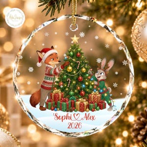 May include: A round, clear acrylic Christmas ornament featuring a printed illustration of a fox and a rabbit decorating a green pine tree with ornaments and lights. Below the tree, the text reads Sophi & Alex and the year 2026 in red script.