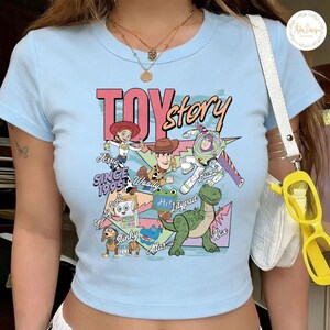 Pode incluir: Uma t-shirt de manga curta em azul claro com uma impress&atilde;o gr&aacute;fica colorida de personagens de Toy Story, incluindo Jessie, Woody, Buzz Lightyear, Rex e Slinky, com o texto Toy Story, Since 1995 e os nomes das personagens em v&aacute;rias fontes.
