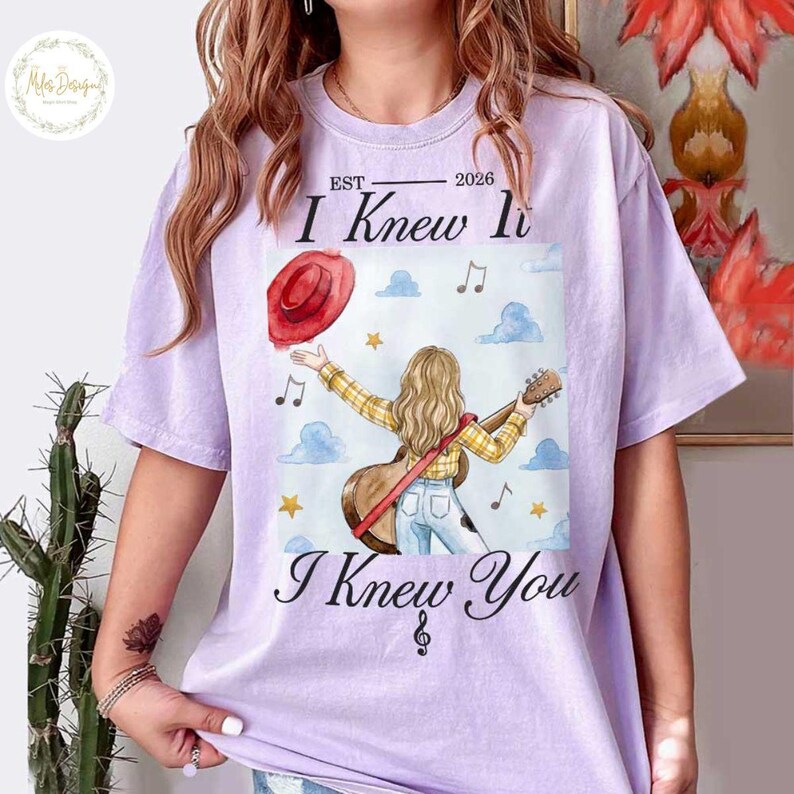 Op de afbeelding: Een lichtpaars t-shirt met een afbeelding van een persoon met lang blond haar die op een akoestische gitaar speelt. Het ontwerp bevat een rode hoed, blauwe wolken, gele sterren, muzieknoten en de tekst EST 2026 I Knew It I Knew You.