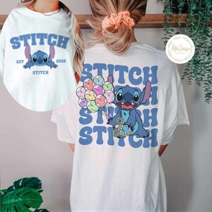 K&ouml;nnte beinhalten: Wei&szlig;es T-Shirt mit einer blauen Stitch-Figur, die auf der R&uuml;ckseite Ballons h&auml;lt. Die Vorderseite des Shirts tr&auml;gt den Text "Stitch Est 2002 Stitch" in Blau.
