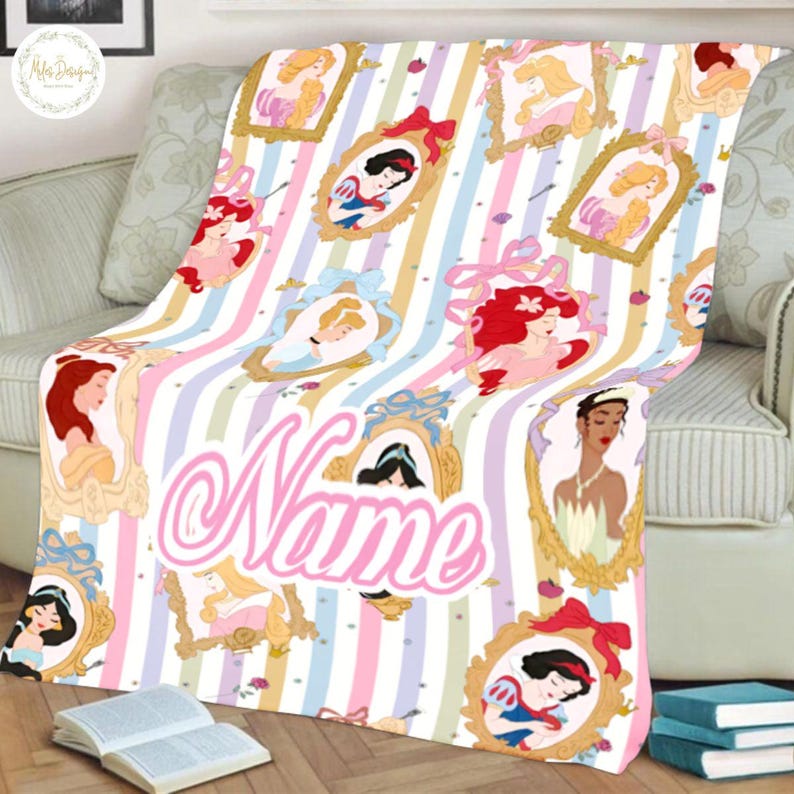 May include: A soft, pastel-striped blanket featuring framed illustrations of various princesses. The blanket has a large, cursive "Name" in pink. The design includes images of Snow White, Ariel, and other Disney princesses. The blanket is draped over a sofa.