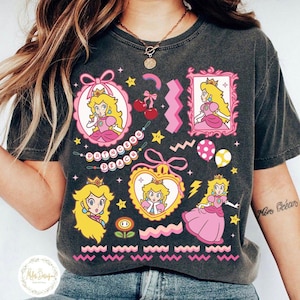 May include: Dark gray t-shirt featuring Princess Peach graphics in pink, yellow, and white. The design includes various Princess Peach portraits, stars, cherries, and the text "Princess Peach". The shirt has a vintage, washed look.