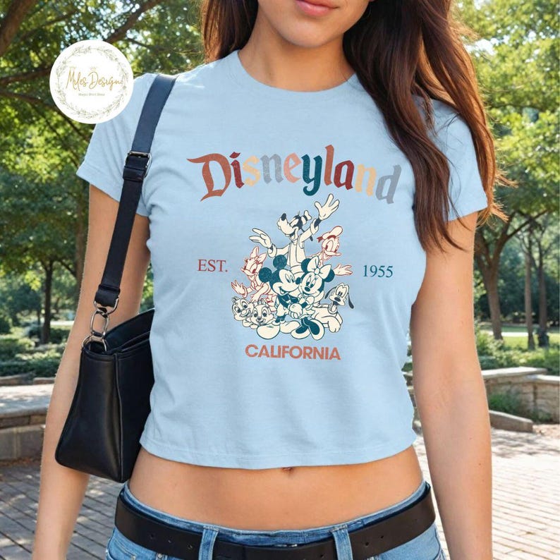 May include: A light blue cropped t-shirt featuring a vintage-style graphic of Mickey Mouse and his friends. The graphic includes the text "Disneyland", "Est. 1955", and "California".