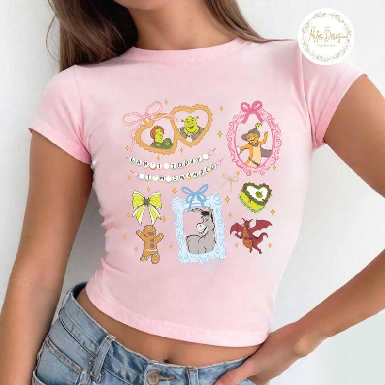 Op de afbeelding: Roze getailleerd T-shirt met een grafische print van personages uit de film Shrek. De grafiek bevat Shrek, Fiona, Ezel, de Kat in Laarzen en de Peperkoekman. De tekst op het shirt luidt "Can't today, I'm swamped."