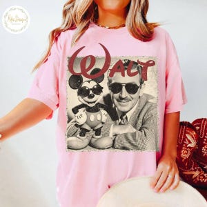 May include: Pale pink t-shirt with a retro graphic of Mickey Mouse and Walt Disney, both sporting sunglasses. The word "Walt" is printed in burgundy above the image. A classic design for Disney fans.