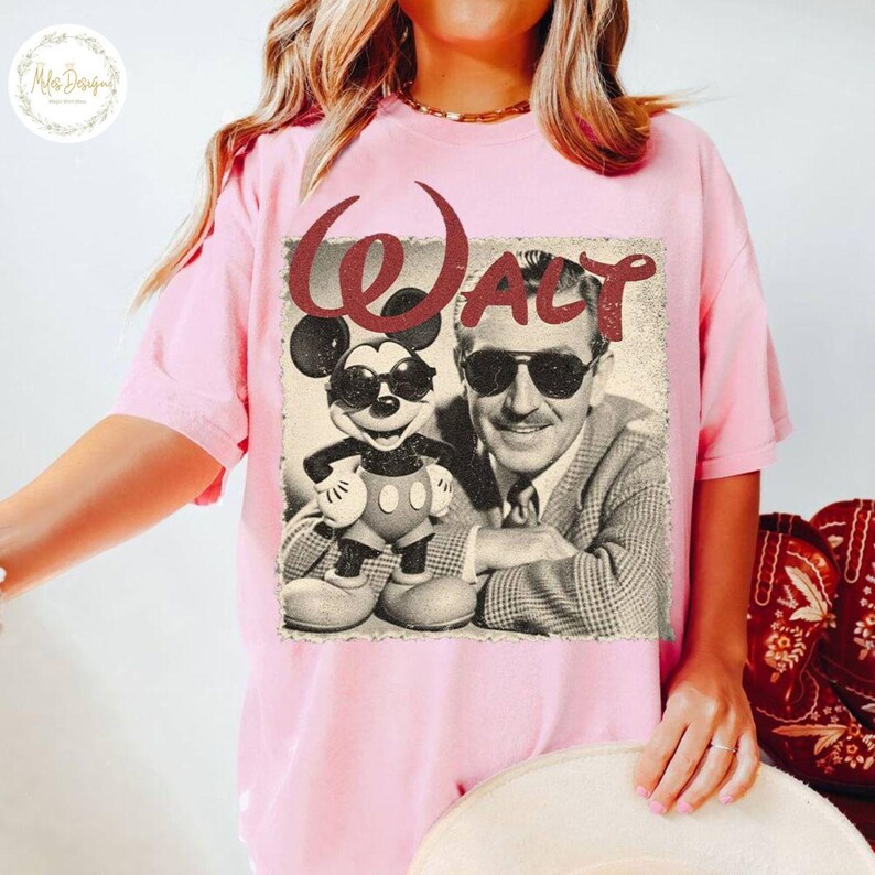 May include: Pale pink t-shirt with a retro graphic of Mickey Mouse and Walt Disney, both sporting sunglasses. The word "Walt" is printed in burgundy above the image. A classic design for Disney fans.