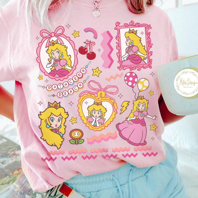 May include: Pink graphic t-shirt featuring Princess Peach from the Super Mario franchise. The design includes multiple framed portraits, stars, cherries, and the text "Princess Peach". The shirt has a crew neck and short sleeves.