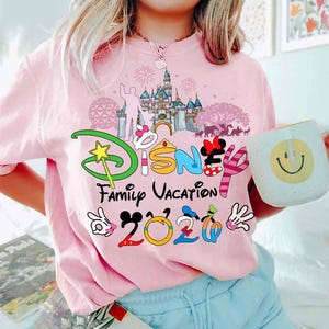 May include: Pink t-shirt with a colorful Disney graphic. The design includes the word "Disney" with Mickey Mouse ears, a castle, and the words "Family Vacation 2020". A white mug with a smiley face is held in the hand.