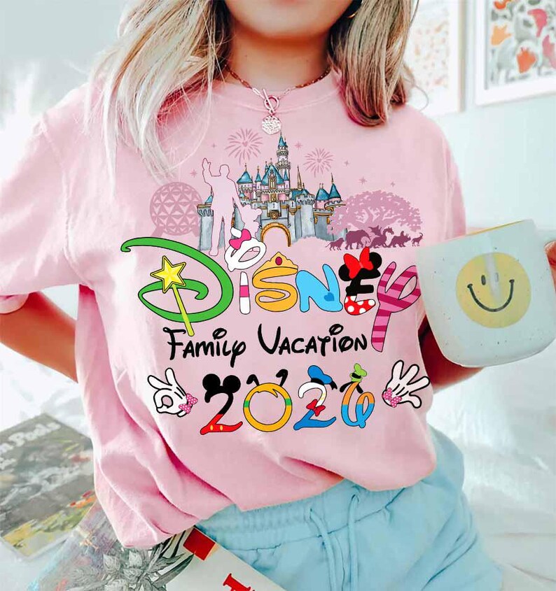 May include: Pink t-shirt with a colorful Disney graphic. The design includes the word "Disney" with Mickey Mouse ears, a castle, and the words "Family Vacation 2020". A white mug with a smiley face is held in the hand.