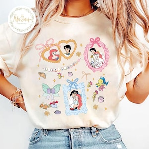Puede incluir: Una camiseta blanca con un dise&ntilde;o colorido que presenta a Ariel y al Pr&iacute;ncipe Eric de La Sirenita. El dise&ntilde;o incluye corazones, conchas marinas y una pancarta que dice "Ariel & Noah".