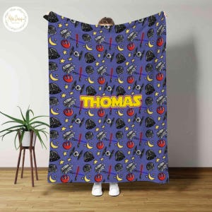 May include: A blue blanket featuring a repeating pattern of Star Wars-themed elements, including Darth Vader helmets, lightsabers, and starships. The name "THOMAS" is prominently displayed in yellow block letters.
