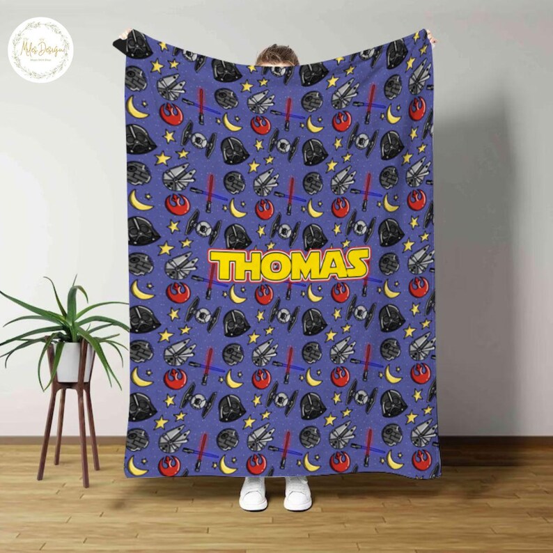 May include: A blue blanket featuring a repeating pattern of Star Wars-themed elements, including Darth Vader helmets, lightsabers, and starships. The name "THOMAS" is prominently displayed in yellow block letters.
