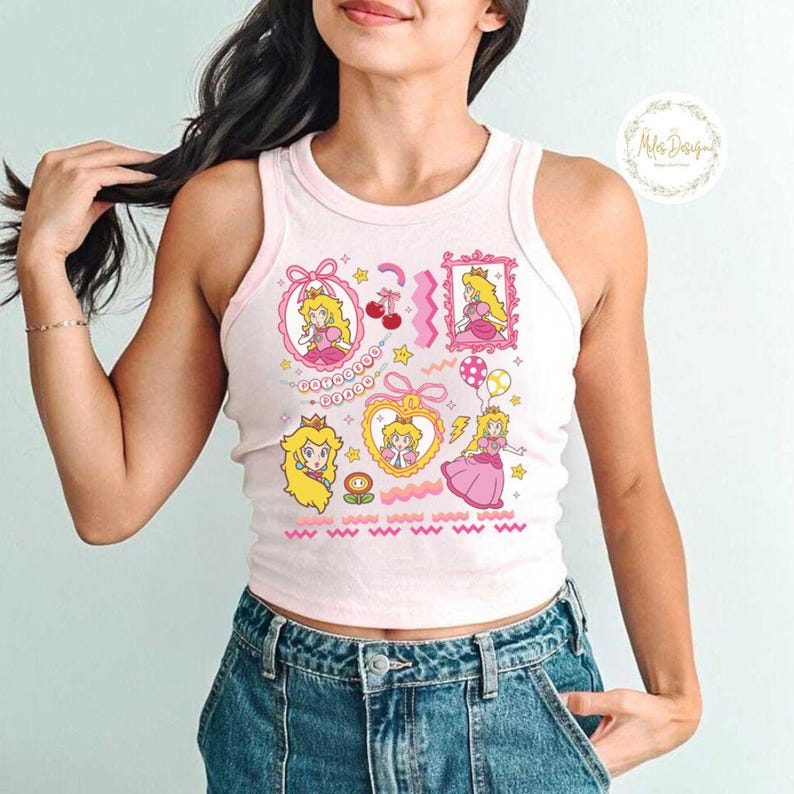 May include: A light pink tank top featuring multiple cartoon illustrations of a princess in a pink dress, with gold crowns, and pink and yellow accents. The tank top has a round neckline and a ribbed texture.