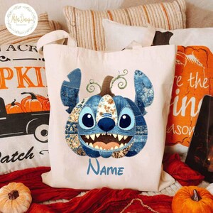 Pode incluir: Uma sacola creme com um design de patchwork azul do rosto de um personagem, que se assemelha a uma ab&oacute;bora. O personagem tem olhos grandes, um sorriso largo e a palavra "Name" em azul. A sacola tem duas al&ccedil;as.