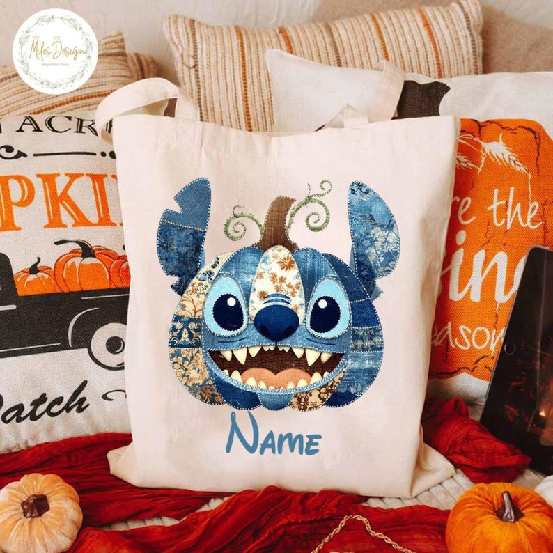 Pode incluir: Uma sacola creme com um design de patchwork azul do rosto de um personagem, que se assemelha a uma ab&oacute;bora. O personagem tem olhos grandes, um sorriso largo e a palavra "Name" em azul. A sacola tem duas al&ccedil;as.