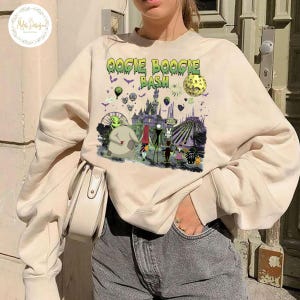 May include: Beige sweatshirt with a colorful graphic featuring the text "Oogie Boogie Bash" and various Halloween-themed characters and elements. The design includes a castle, a full moon, and other spooky details.