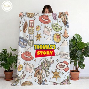 May include: A light blue blanket featuring cartoon characters and elements from the movie Toy Story. The blanket includes the text "THOMAS'S STORY" in a red and yellow design, along with images of Woody, Buzz Lightyear, and other characters.
