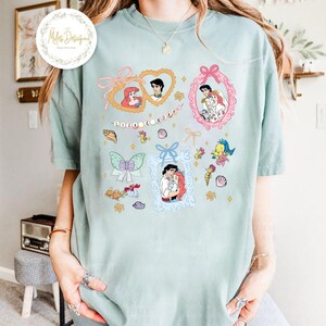 Puede incluir: Una camiseta azul claro con un estampado gr&aacute;fico de Ariel y Eric de La Sirenita. El gr&aacute;fico incluye varios marcos con diferentes escenas de la pel&iacute;cula, incluyendo un marco en forma de coraz&oacute;n con Ariel y Eric bes&aacute;ndose, un marco con Ariel y Eric cogidos de la mano y un marco con Ariel y Eric como pareja. El gr&aacute;fico tambi&eacute;n incluye una cadena de perlas con las palabras "Ariel & Eric" escritas en cuentas.