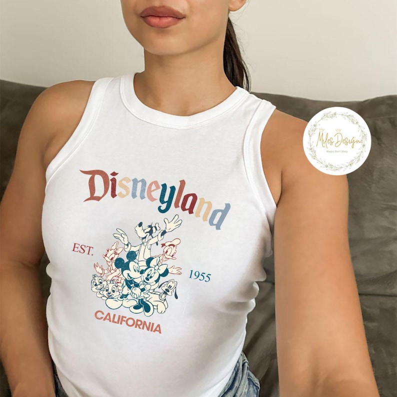 May include: White tank top with a colourful Disneyland graphic featuring Mickey Mouse and friends. The graphic includes the text "Disneyland", "EST. 1955", and "CALIFORNIA".