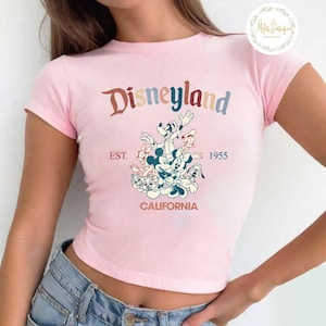May include: Pink cropped t-shirt featuring a vintage-style graphic of Mickey Mouse and friends in front of a Disneyland logo. The text reads "Disneyland EST. 1955 CALIFORNIA".