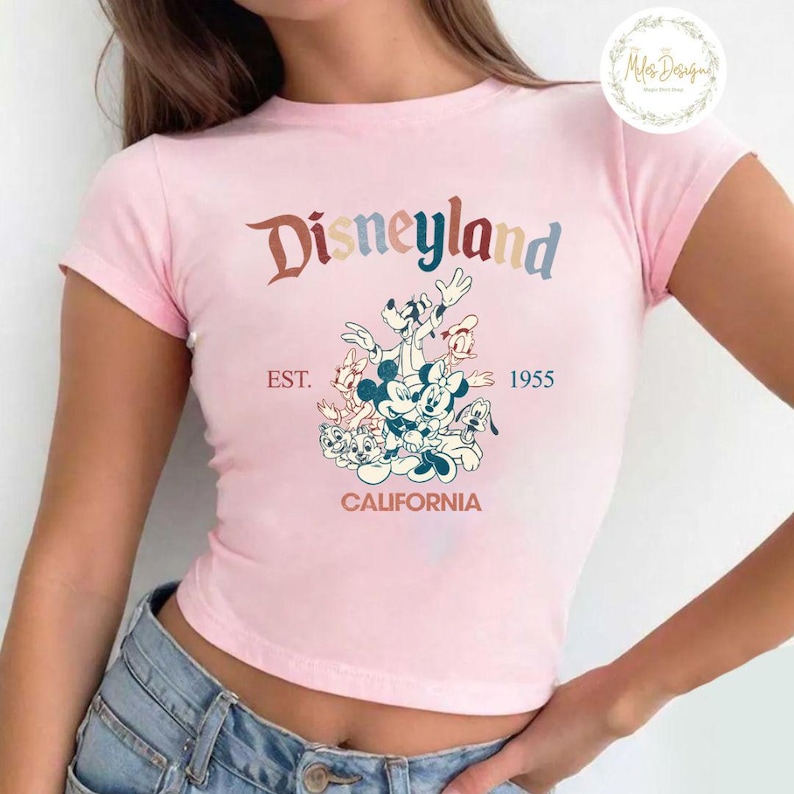 May include: Pink cropped t-shirt featuring a vintage-style graphic of Mickey Mouse and friends in front of a Disneyland logo. The text reads "Disneyland EST. 1955 CALIFORNIA".