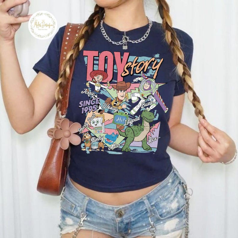 Pode incluir: Uma t-shirt azul-marinho de manga curta com uma estampa gr&aacute;fica colorida de Toy Story. O design inclui as personagens Jessie, Woody, Buzz Lightyear, Slinky Dog, Rex e outras com o texto Toy Story, Since 1995 e nomes das personagens.