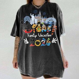 May include: Dark gray t-shirt featuring a colorful Disney design. The shirt includes the text "Disney Family Vacation 2020" with images of Disney characters and a castle silhouette. The shirt has short sleeves and a relaxed fit.