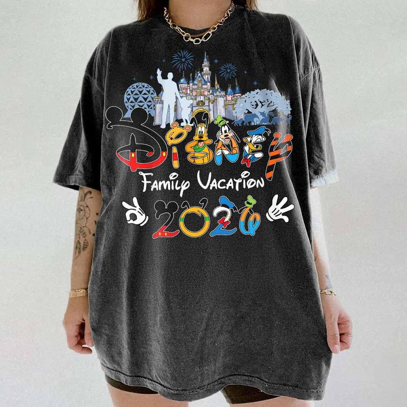 May include: Dark gray t-shirt featuring a colorful Disney design. The shirt includes the text "Disney Family Vacation 2020" with images of Disney characters and a castle silhouette. The shirt has short sleeves and a relaxed fit.