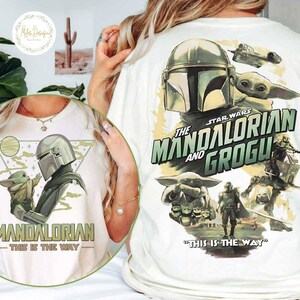 Peut inclure: T-shirt blanc avec un motif de The Mandalorian et Grogu de Star Wars. Le design comprend le texte "The Mandalorian and Grogu" et "This is the Way". Le t-shirt a un col rond et des manches courtes.