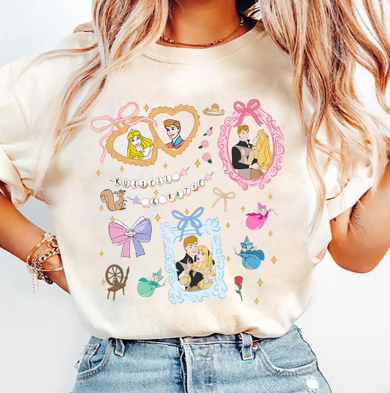 Op de afbeelding: Een wit T-shirt met een kleurrijke grafische print met een cartoon afbeelding van een stel, een eekhoorn, een spinnewiel, een strik en een fee. De tekst "RUSSELL & KATIE" is gedrukt in een kleurrijk lettertype.
