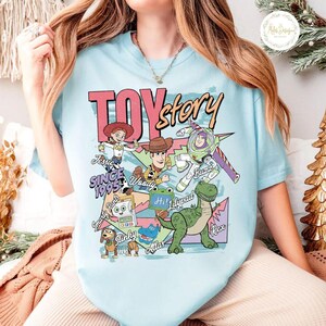 Pode incluir: Uma t-shirt azul-clara com uma impress&atilde;o gr&aacute;fica colorida das personagens de Toy Story, incluindo Woody, Buzz Lightyear, Jessie, Rex, Slinky Dog e Smarty Pants. O design inclui o texto Toy Story, Since 1995 e os nomes das personagens.
