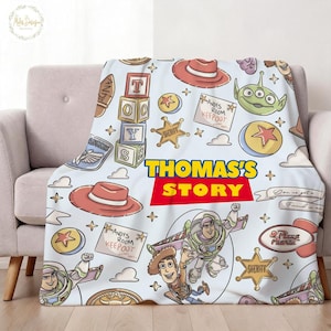 May include: A light blue blanket featuring cartoon characters and elements from the movie "Toy Story." The design includes Woody, Buzz Lightyear, and other iconic imagery, with the text "THOMAS'S STORY" in a bold, colorful font. The blanket is draped over a light-colored sofa.