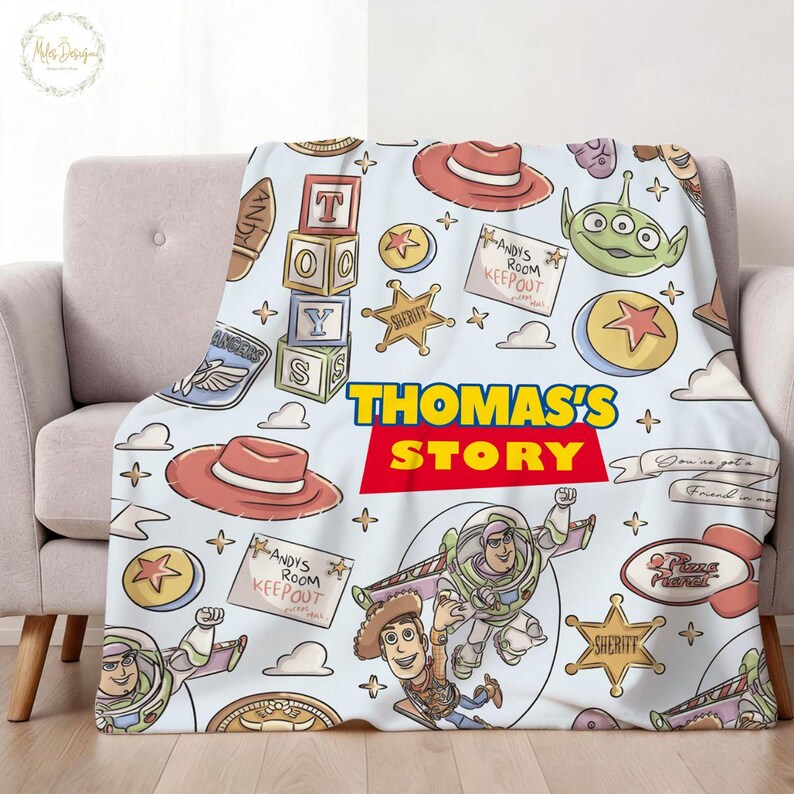 May include: A light blue blanket featuring cartoon characters and elements from the movie "Toy Story." The design includes Woody, Buzz Lightyear, and other iconic imagery, with the text "THOMAS'S STORY" in a bold, colorful font. The blanket is draped over a light-colored sofa.