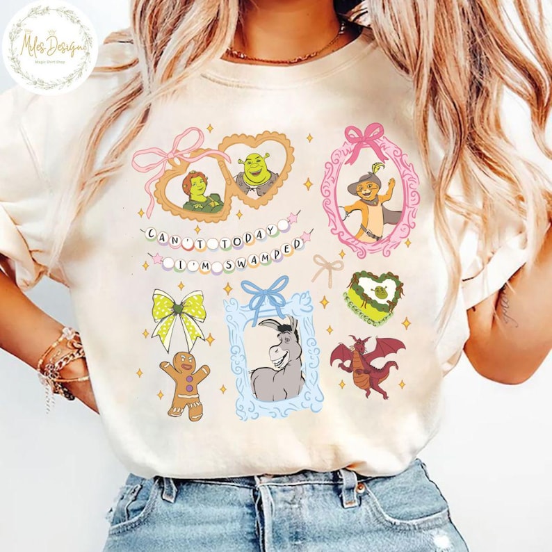 Op de afbeelding: Een wit T-shirt met een grafische print van personages uit de film Shrek. De grafiek bevat Shrek, Fiona, Ezel, Puss in Boots en een gingerbread man. De tekst "Can't today I'm swamped" is op het shirt gedrukt.