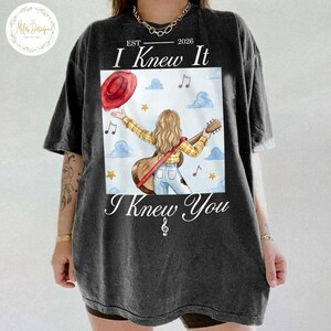 Op de afbeelding: Een antracietgrijs t-shirt met een afbeelding van een persoon met lang blond haar in een geel geruit overhemd en een blauwe spijkerbroek, met een akoestische gitaar. Boven de afbeelding staat EST 2026 I Knew It en onderaan I Knew You.