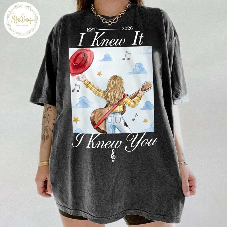 Op de afbeelding: Een antracietgrijs t-shirt met een afbeelding van een persoon met lang blond haar in een geel geruit overhemd en een blauwe spijkerbroek, met een akoestische gitaar. Boven de afbeelding staat EST 2026 I Knew It en onderaan I Knew You.