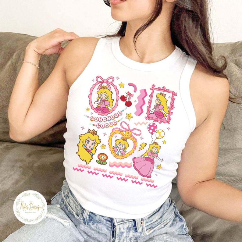May include: White tank top featuring a colorful cartoon graphic of Princess Peach from the Super Mario video game series. The design includes various framed images, stars, cherries, and wavy lines in pink, yellow, and gold.
